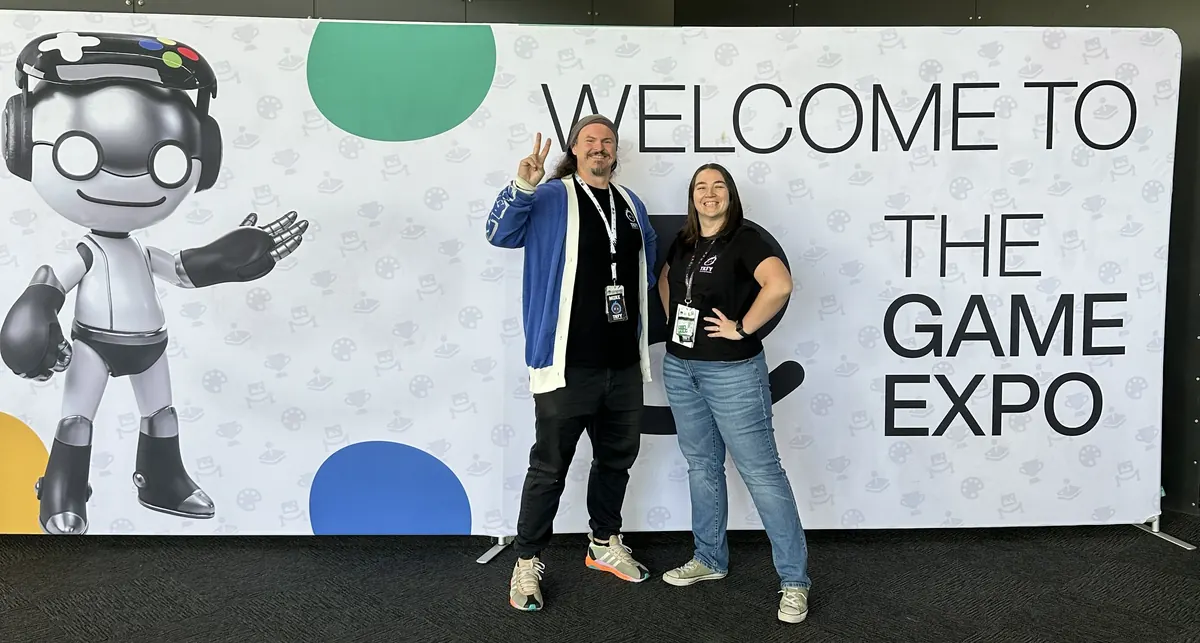 Mike, throwing a peace sign, and Tahnee in TKFY shirts and lanyards in front of a Welcome to The Game Expo backdrop.