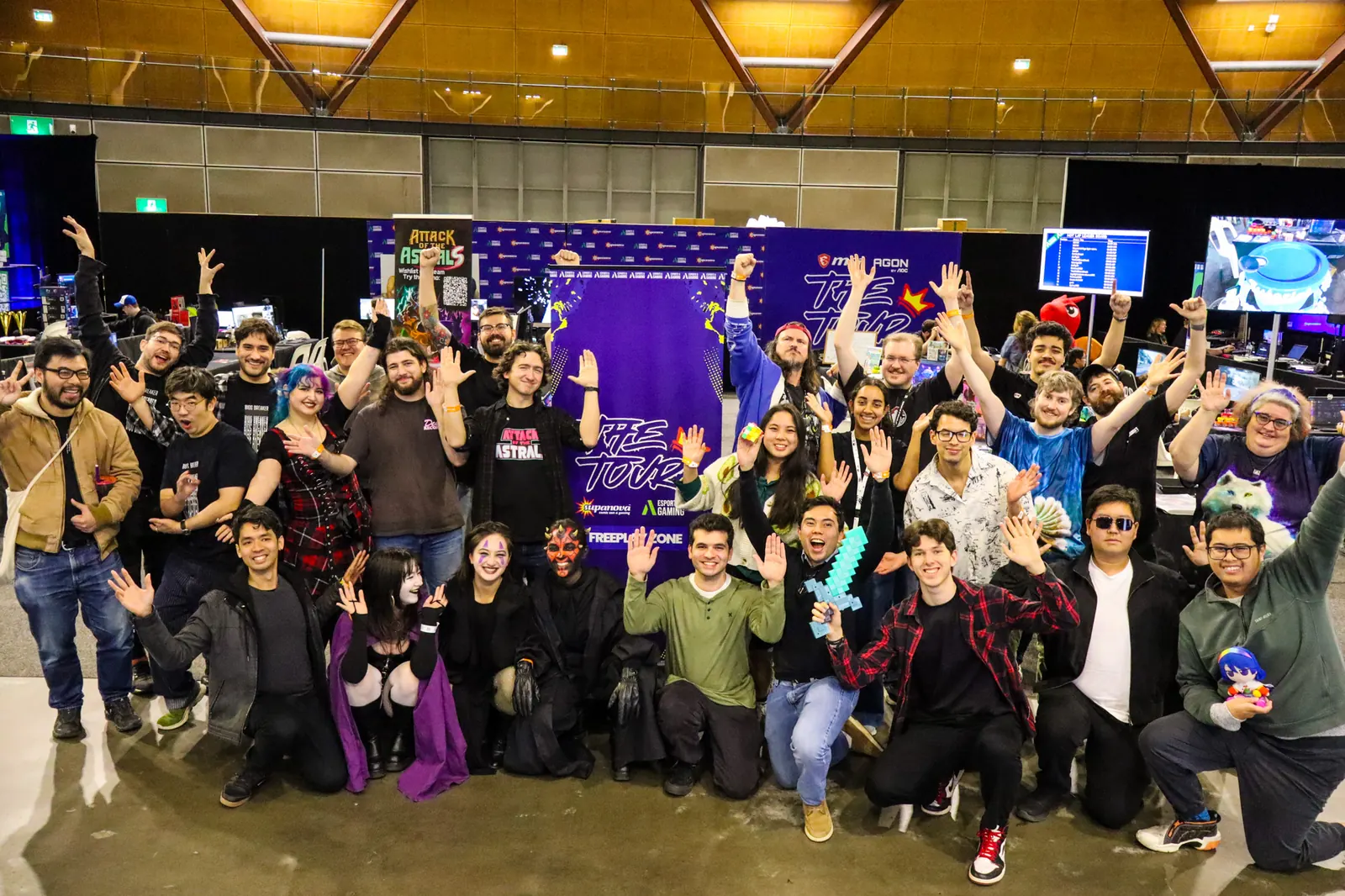 Around forty NSW indie developers and cosplayers pose with arms raised in front of a Freeplay Zone banner at Supanova Sydney. Mike stands in the back row in a red cap and blue cardigan, fist in the air.